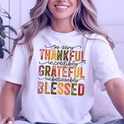 May include: A white short-sleeved t-shirt featuring the text "so very THANKFUL incredibly GRATEFUL unbelievably BLESSED" printed in a mix of black cursive script and block letters in shades of orange, yellow, brown, and dark green.