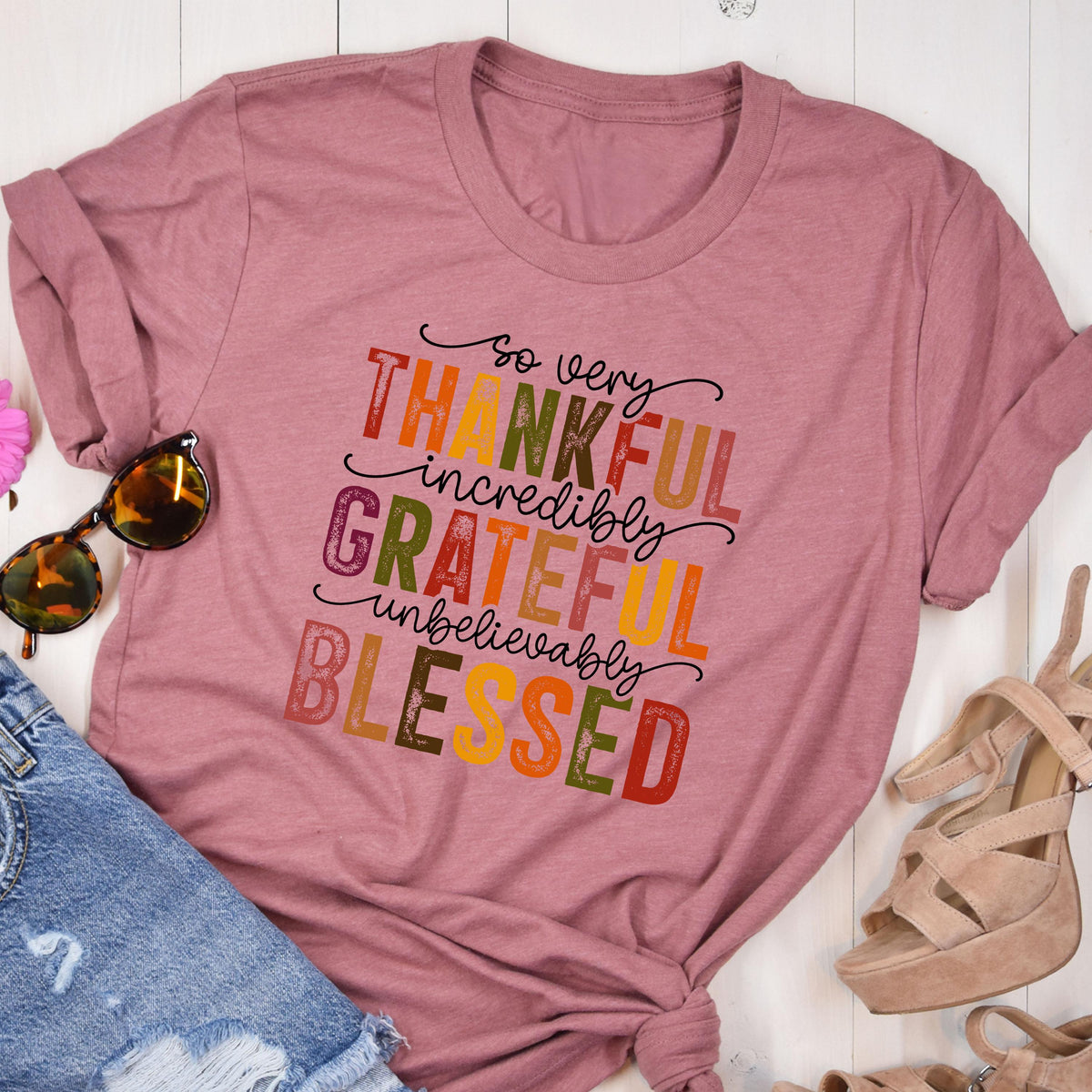 May include: A mauve short-sleeved t-shirt featuring the text "so very THANKFUL incredibly GRATEFUL unbelievably BLESSED" in a multi-colored, distressed font. The shirt is displayed alongside a pair of sunglasses and beige heeled sandals.
