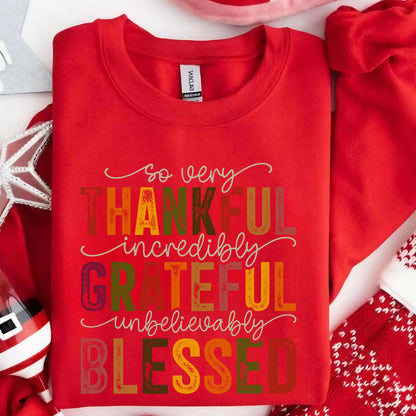 May include: A bright red crewneck sweatshirt featuring the text "so very THANKFUL incredibly GRATEFUL unbelievably BLESSED" printed in a mix of autumn-toned colors including orange, yellow, green, and brown, with a white tag at the neckline.