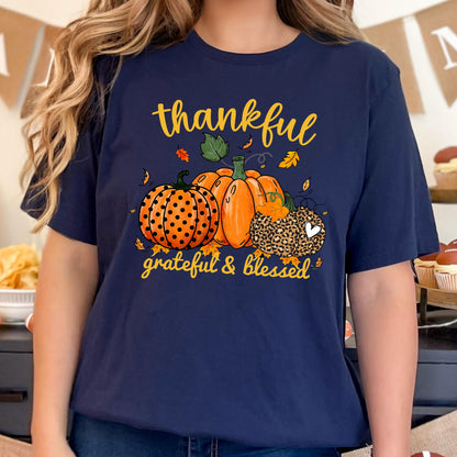 May include: Navy blue t-shirt featuring a fall-themed graphic. The design includes three pumpkins in orange and brown, with the words "thankful, grateful & blessed" in yellow script. The pumpkins have polka dots and leopard print.