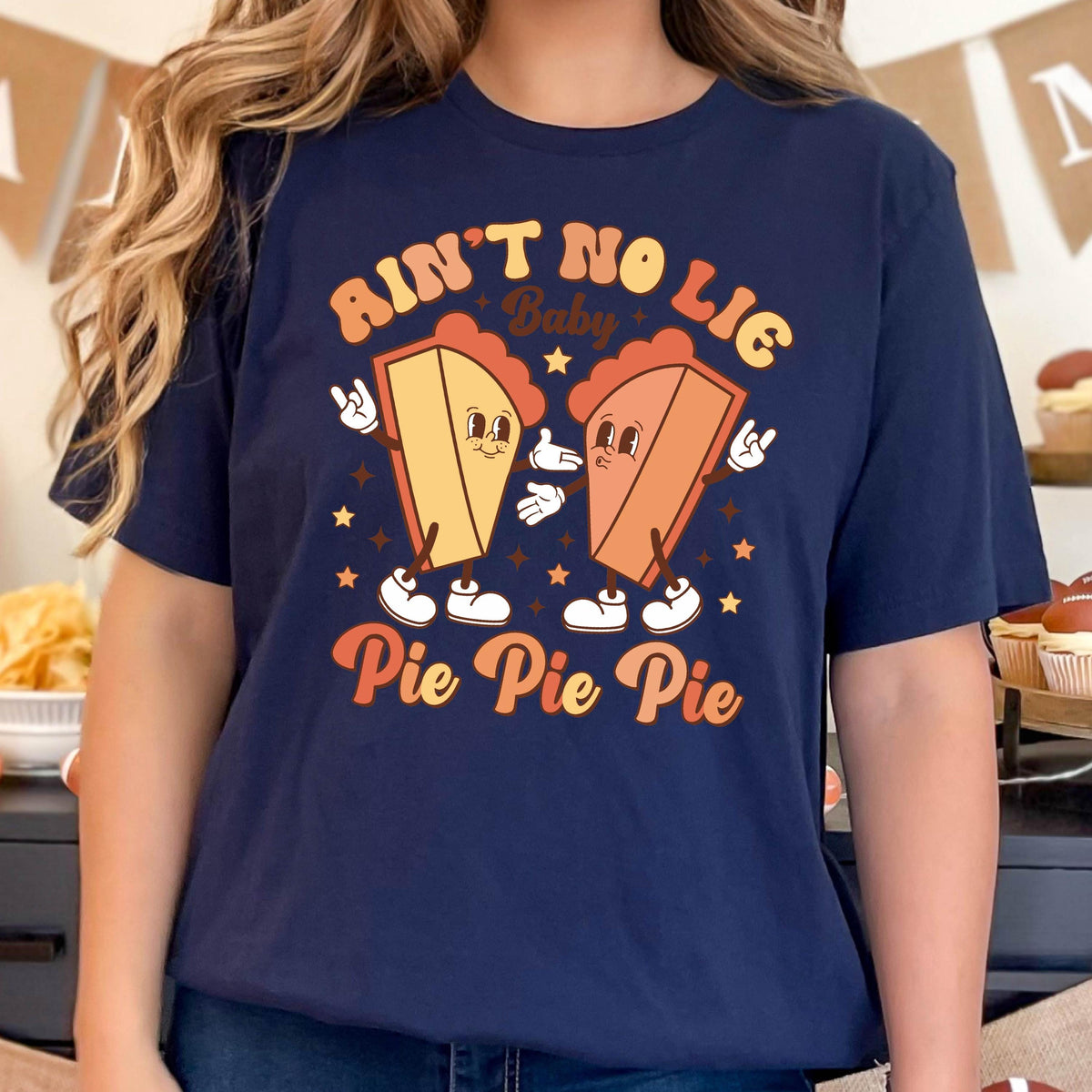 May include: Navy blue t-shirt with a retro design featuring two cartoon pie slices with arms and legs. The text reads "Ain't No Lie Baby" above and "Pie Pie Pie" below the pie characters. Stars are scattered around the design.