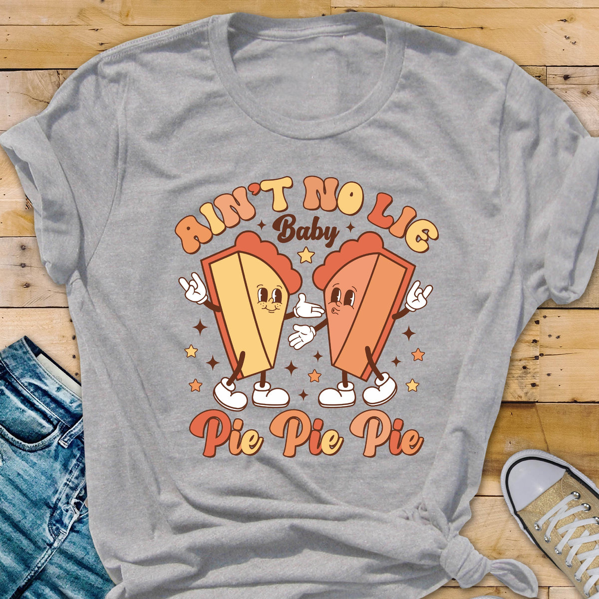 May include: A gray t-shirt featuring a retro design with the text "Ain't No Lie Baby" above two cartoon pie slices. Below the text is "Pie Pie Pie". The pie slices are orange and yellow with white shoes and star accents.