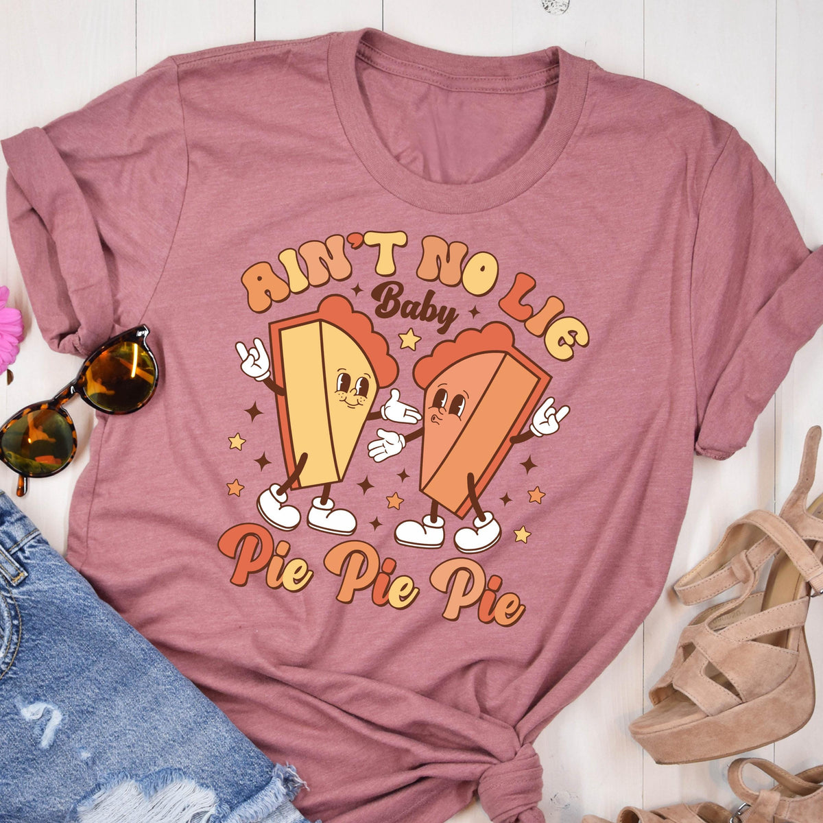 May include: Mauve t-shirt featuring a retro graphic of two pie slices with faces, arms, and legs. The text "Ain't No Lie Baby" and "Pie Pie Pie" are in a vintage font. Sunglasses and denim shorts are visible.