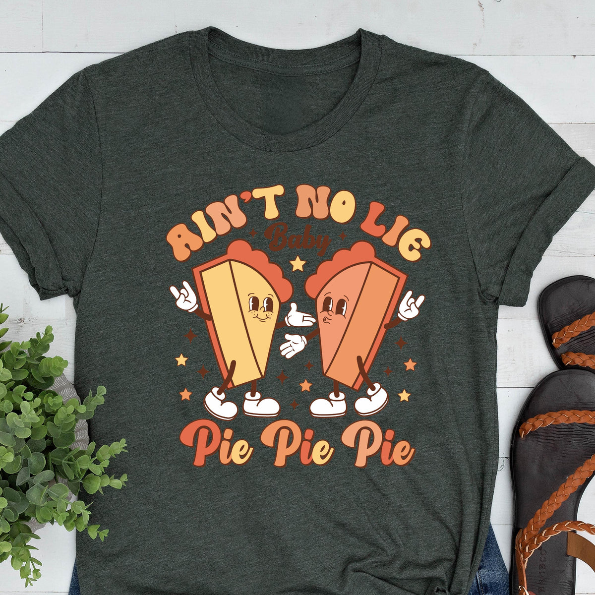May include: Dark green t-shirt featuring a retro graphic of two pie slices with arms, legs, and faces. The text on the shirt reads "Ain't No Lie Baby Pie Pie Pie" in orange and brown lettering. The design includes stars and a vintage aesthetic.