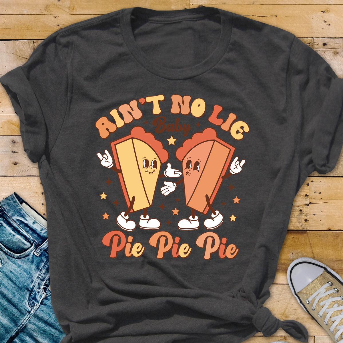 May include: Dark gray t-shirt with a retro graphic design. The design features two cartoon pie slices with arms, legs, and faces, with the text "Ain't No Lie Baby" above and "Pie Pie Pie" below. The pies are in shades of orange and yellow.