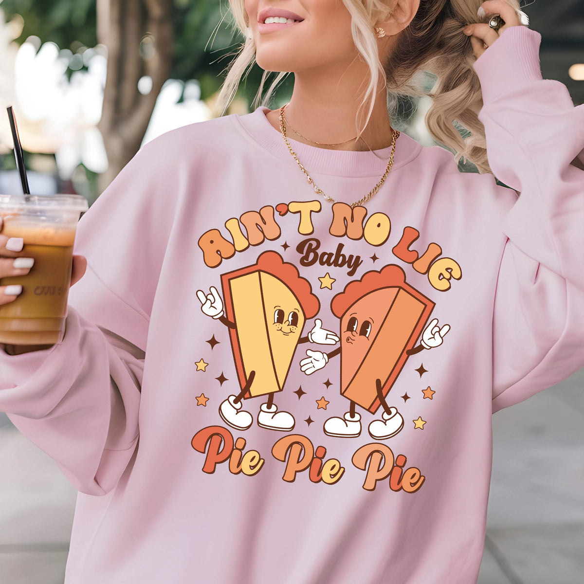 May include: A light pink crewneck sweatshirt featuring a graphic print of two cartoon pie slices with arms and legs. The text reads "AIN'T NO LIE Baby Pie Pie Pie" in a retro font, surrounded by small star shapes in orange and yellow tones.