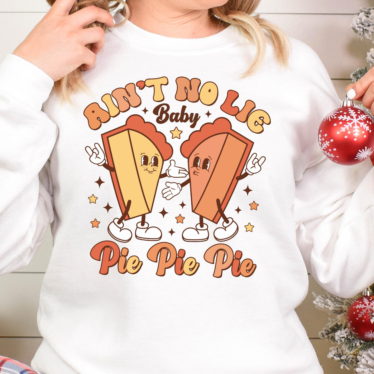 May include: A white long-sleeve sweatshirt featuring a graphic of two cartoon pie slices with arms and legs. The text reads "AIN'T NO LIE Baby" at the top and "Pie Pie Pie" at the bottom in orange and yellow retro-style lettering with stars.