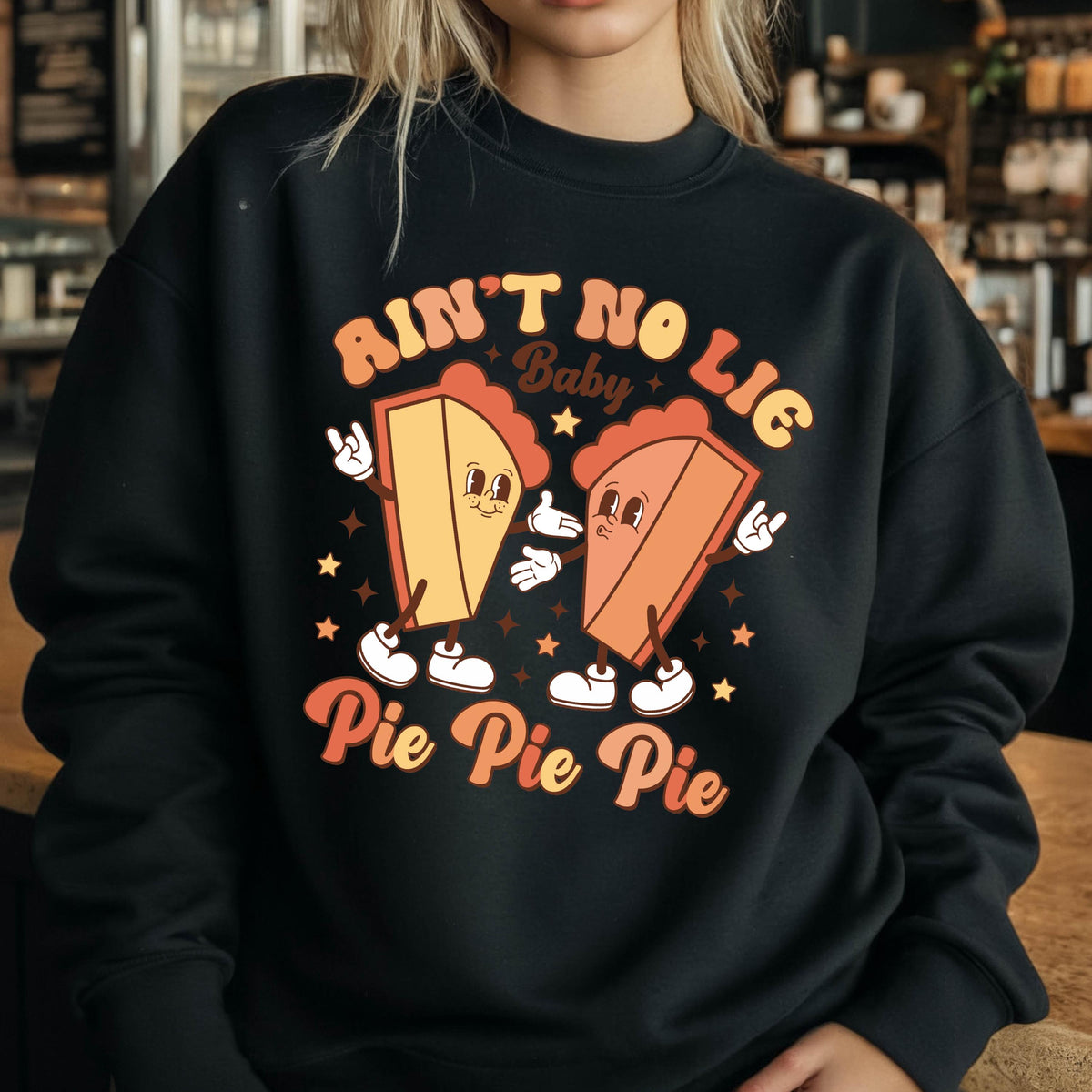 May include: A black crewneck sweatshirt featuring a graphic print of two anthropomorphic pie slices with arms and legs. The text reads "AIN'T NO LIE Baby Pie Pie Pie" in a retro font, surrounded by small stars in shades of orange, yellow, and cream.