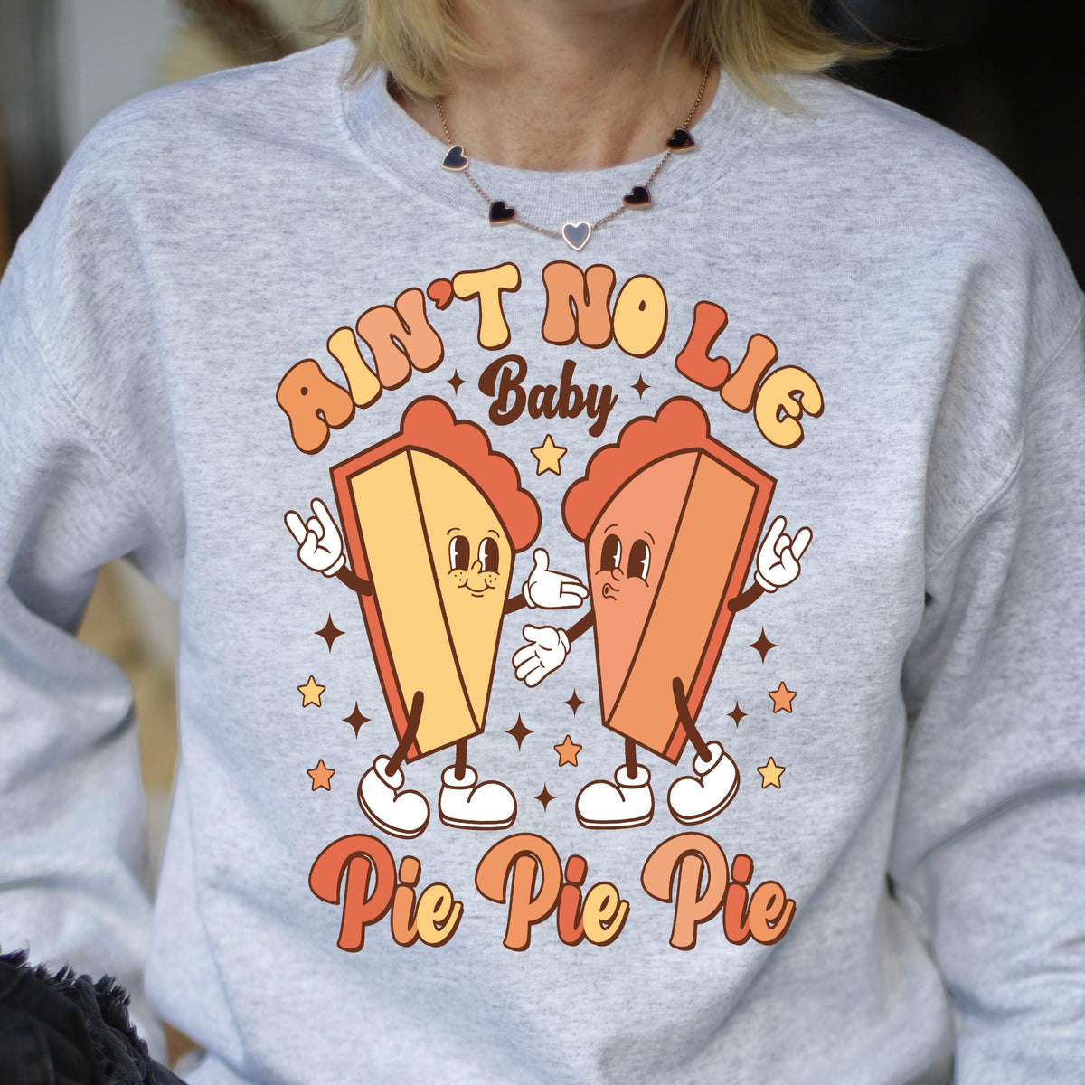 May include: A light gray crewneck sweatshirt featuring a graphic print of two anthropomorphic pie slices with arms and legs. The text reads AIN'T NO LIE Baby Pie Pie Pie. The pie slices are yellow and orange with white gloves and shoes.
