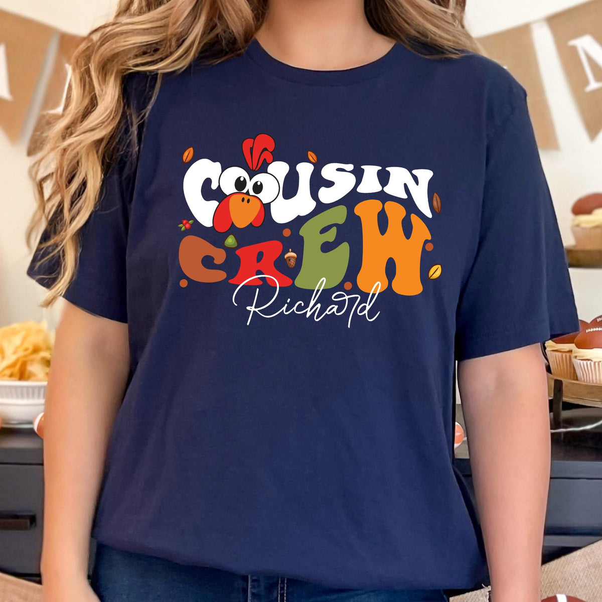 May include: A navy blue short-sleeve t-shirt featuring a graphic print with the text "COUSIN CREW" in large, colorful, retro-style lettering. A cartoon turkey head is integrated into the design, along with the name "Richard" in white script.