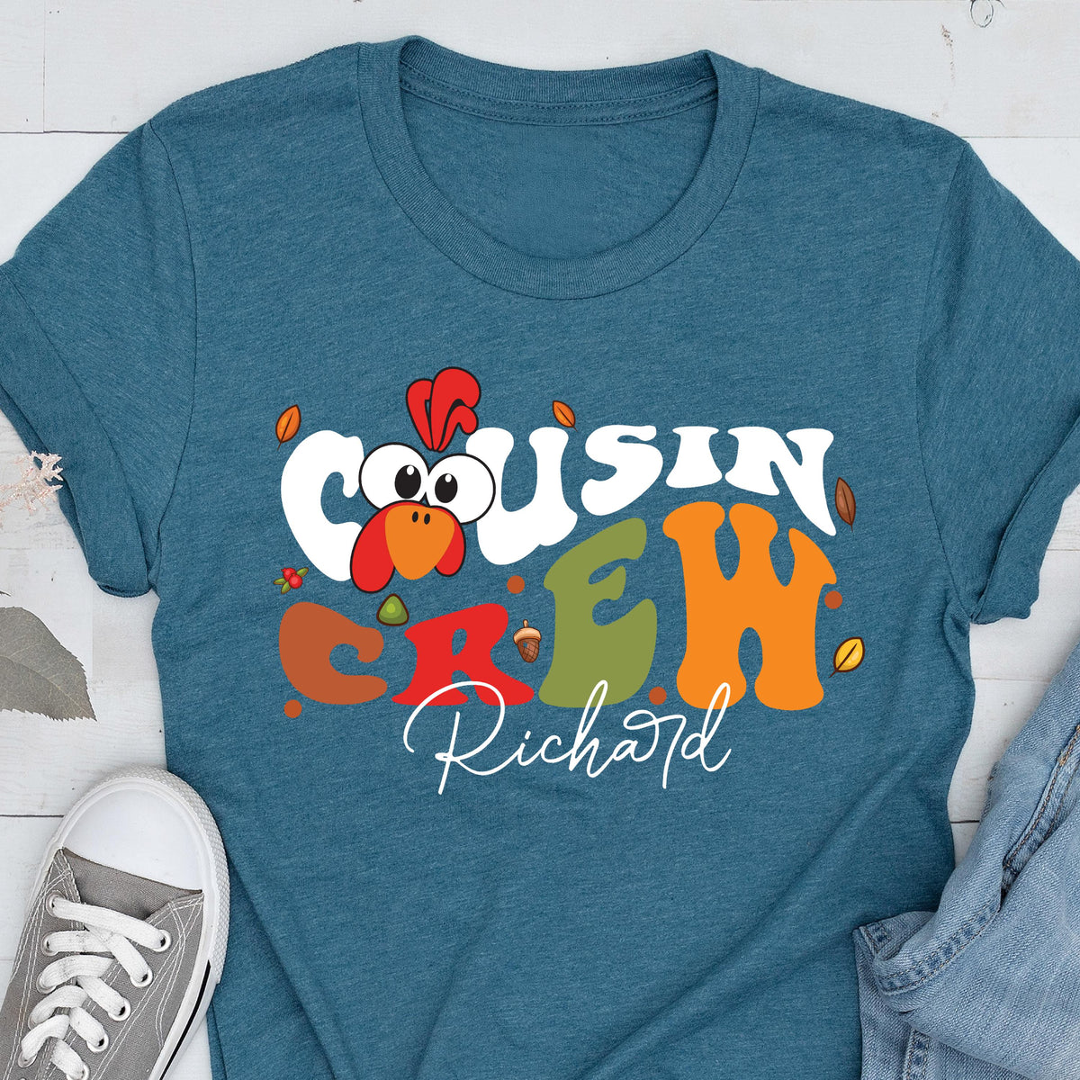 May include: A teal short-sleeve t-shirt featuring a graphic of a cartoon turkey head and the text "COUSIN CREW" in large, colorful, wavy letters. Below the text, the name "Richard" is printed in a white script font, surrounded by small leaves.