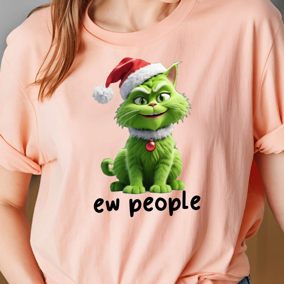 May include: A peach-colored t-shirt featuring a graphic of a green cat wearing a red and white Santa hat and a red ornament collar. The cat has a mischievous expression, and the text "ew people" is printed in black letters below the cat.