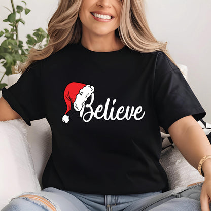 May include: A black short-sleeved t-shirt featuring the word Believe in white cursive script. The letter B is replaced by a red and white Santa Claus hat with a white pom-pom. The design is centered on the chest of the garment.