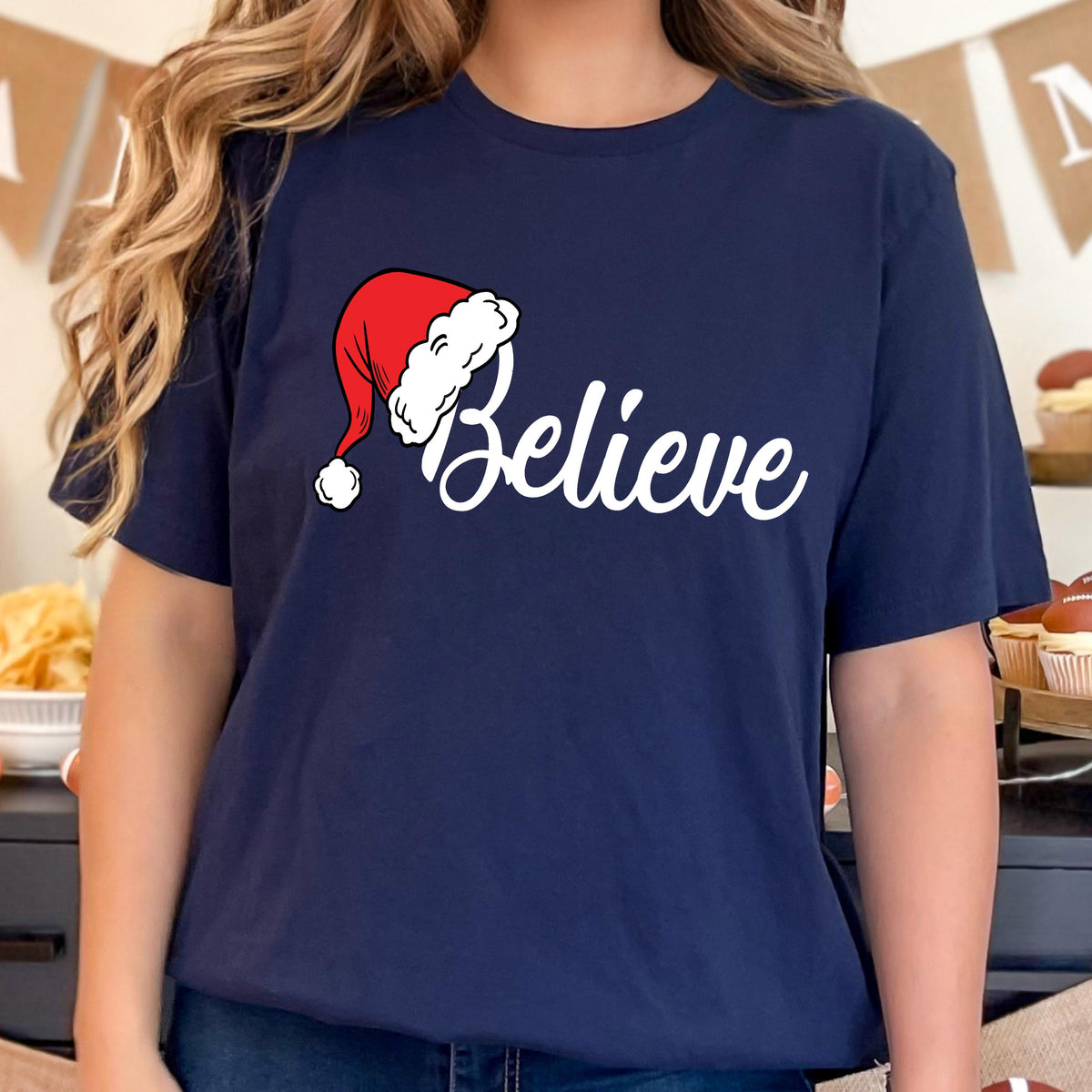 May include: A navy blue short-sleeve t-shirt featuring the white text "Believe" in a cursive script font. The letter "B" is replaced by a red and white Santa Claus hat with a white pom-pom, positioned above the word on the chest area.