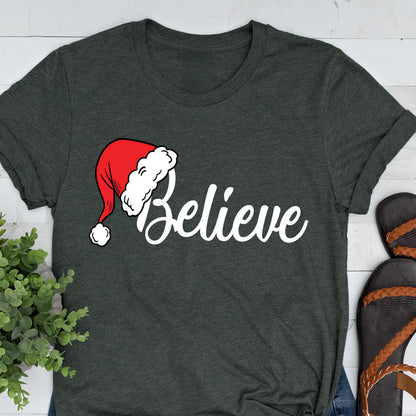 May include: A dark charcoal gray short-sleeve t-shirt featuring the white cursive text "Believe" across the chest. The letter "B" is replaced by a red and white Santa Claus hat with a white pom-pom at the tip, printed on the front of the shirt.