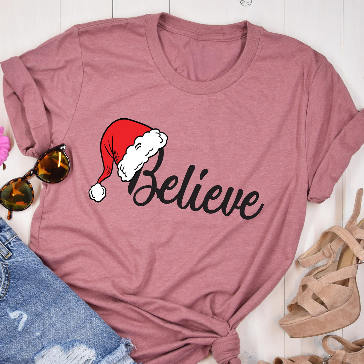 May include: A mauve short-sleeved t-shirt featuring the word Believe in black cursive script. The letter B is topped with a red and white Santa Claus hat. The shirt is displayed alongside a pair of sunglasses, blue denim jeans, and tan heels.