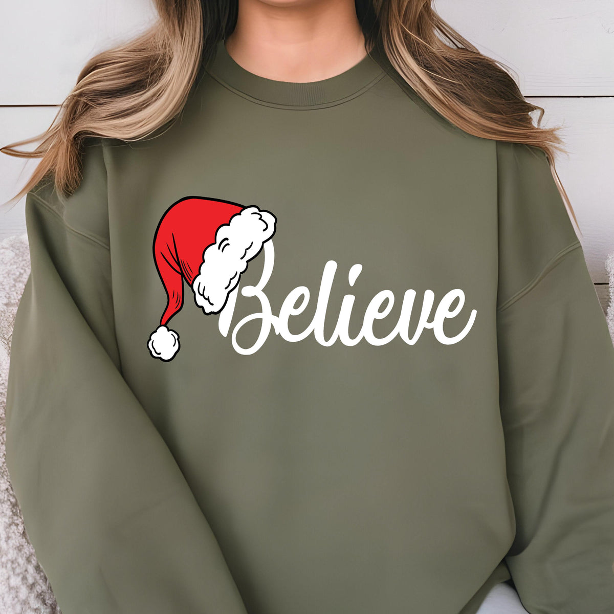 May include: An olive green crewneck sweatshirt featuring a graphic print on the chest. The design includes a red and white Santa hat positioned above the white cursive text that reads Believe, with the hat replacing the letter B in the word.