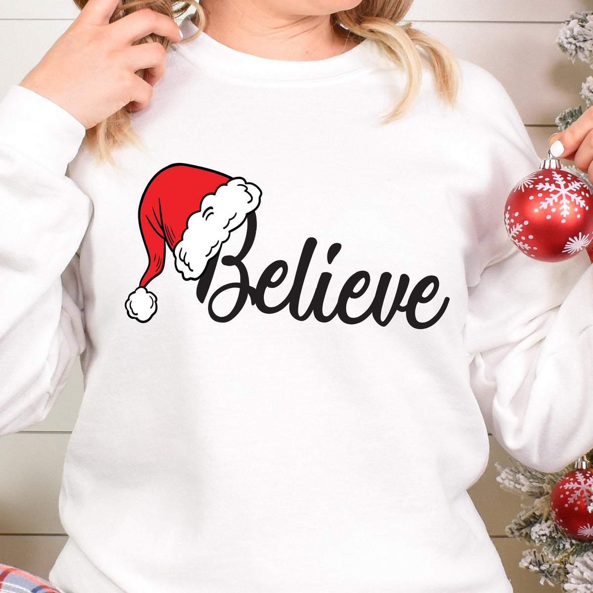 May include: A white long-sleeve sweatshirt featuring a graphic print of a red Santa hat with a white pom-pom and trim, positioned above the black cursive text "Believe". The design is centered on the chest of the garment.