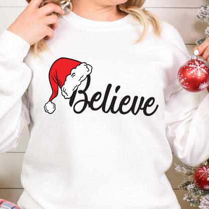 May include: A white long-sleeve sweatshirt featuring a graphic print of a red Santa hat with a white pom-pom and trim, positioned above the black cursive text "Believe". The design is centered on the chest of the garment.