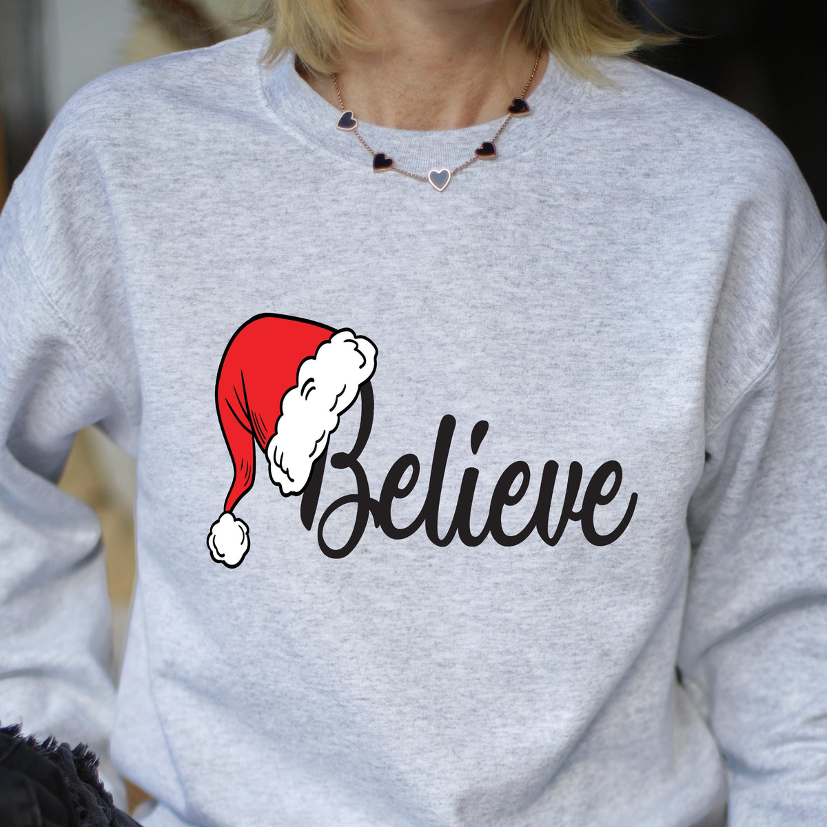 May include: A light gray crewneck sweatshirt featuring a graphic of a red and white Santa hat positioned over the black cursive text "Believe". The garment has long sleeves and a ribbed neckline, providing a simple and clean aesthetic.