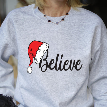 May include: A light gray crewneck sweatshirt featuring a graphic of a red and white Santa hat positioned over the black cursive text "Believe". The garment has long sleeves and a ribbed neckline, providing a simple and clean aesthetic.