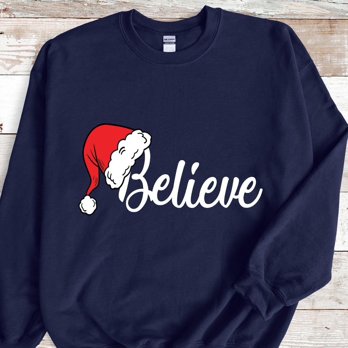 May include: A navy blue long-sleeve sweatshirt featuring the word Believe in white cursive script. The letter B is replaced by a red Santa hat with a white fur trim and a white pom-pom. The garment has a crew neckline and long sleeves.
