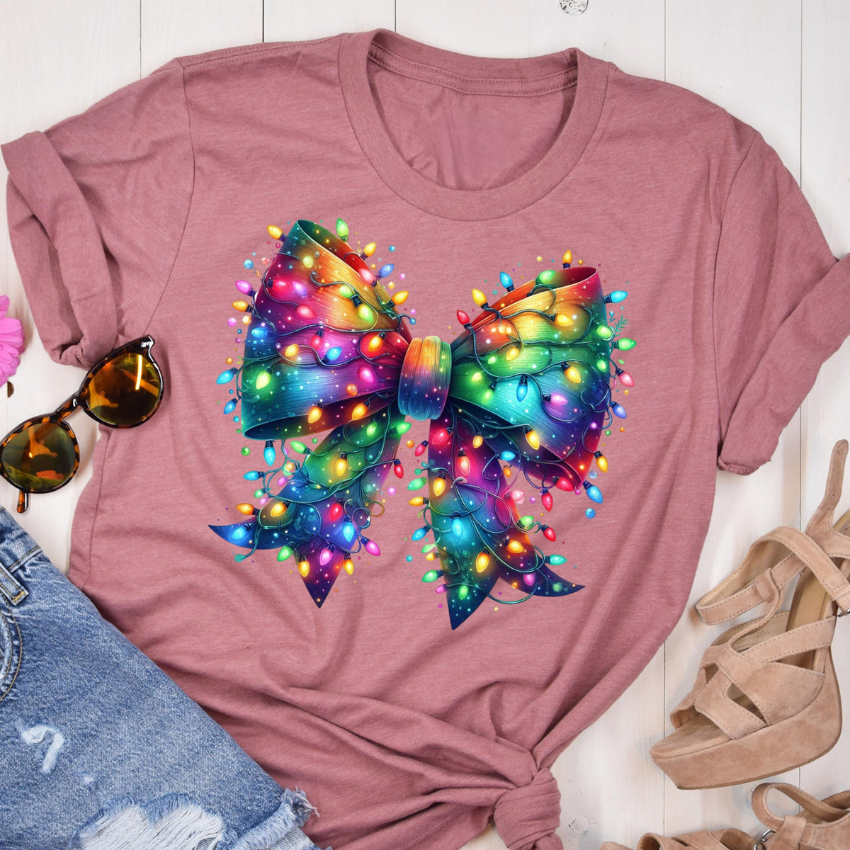 May include: Dusty rose t-shirt featuring a large, colorful bow design. The bow is adorned with strings of vibrant, multicolored Christmas lights. The design is centered on the shirt, creating a festive and eye-catching graphic.