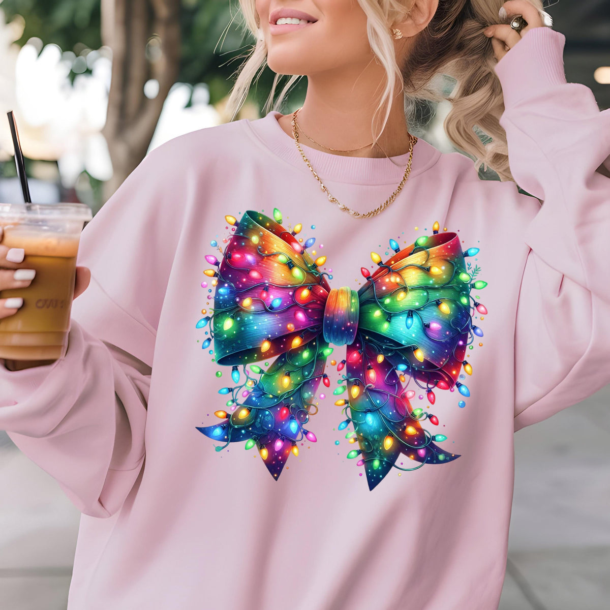 May include: A light pink long-sleeve sweatshirt featuring a large, central graphic of a decorative bow wrapped in vibrant, multi-colored glowing Christmas string lights. The bow displays a spectrum of rainbow colors, including blue, purple, and green.