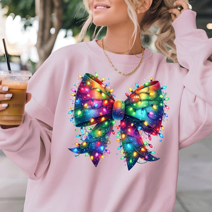 May include: A light pink long-sleeve sweatshirt featuring a large, central graphic of a decorative bow wrapped in vibrant, multi-colored glowing Christmas string lights. The bow displays a spectrum of rainbow colors, including blue, purple, and green.