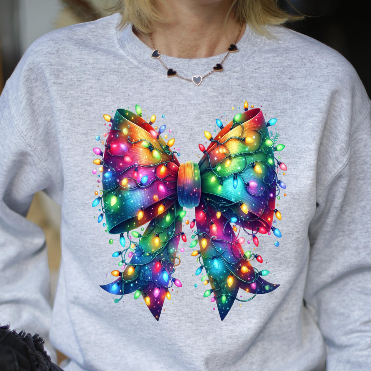 May include: A light gray crewneck sweatshirt featuring a large, central graphic of a vibrant, rainbow-colored bow. The bow is intricately wrapped in glowing, multi-colored string lights in shades of red, blue, yellow, green, and pink.