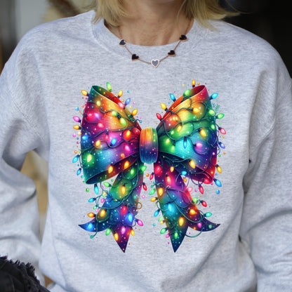 May include: A light gray crewneck sweatshirt featuring a large, central graphic of a vibrant, rainbow-colored bow. The bow is intricately wrapped in glowing, multi-colored string lights in shades of red, blue, yellow, green, and pink.