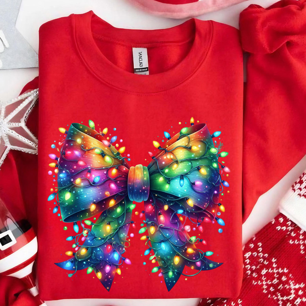 May include: A bright red crewneck sweatshirt featuring a large, central graphic of a bow made from colorful, glowing Christmas string lights. The bow displays a vibrant rainbow gradient of colors, with individual bulbs in blue, yellow, and green.