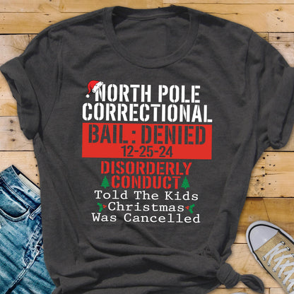 May include: A dark gray short-sleeve t-shirt featuring white and red text that reads North Pole Correctional, Bail: Denied, 12-25-24, Disorderly Conduct, Told The Kids Christmas Was Cancelled, with a small red Santa hat graphic above the text.
