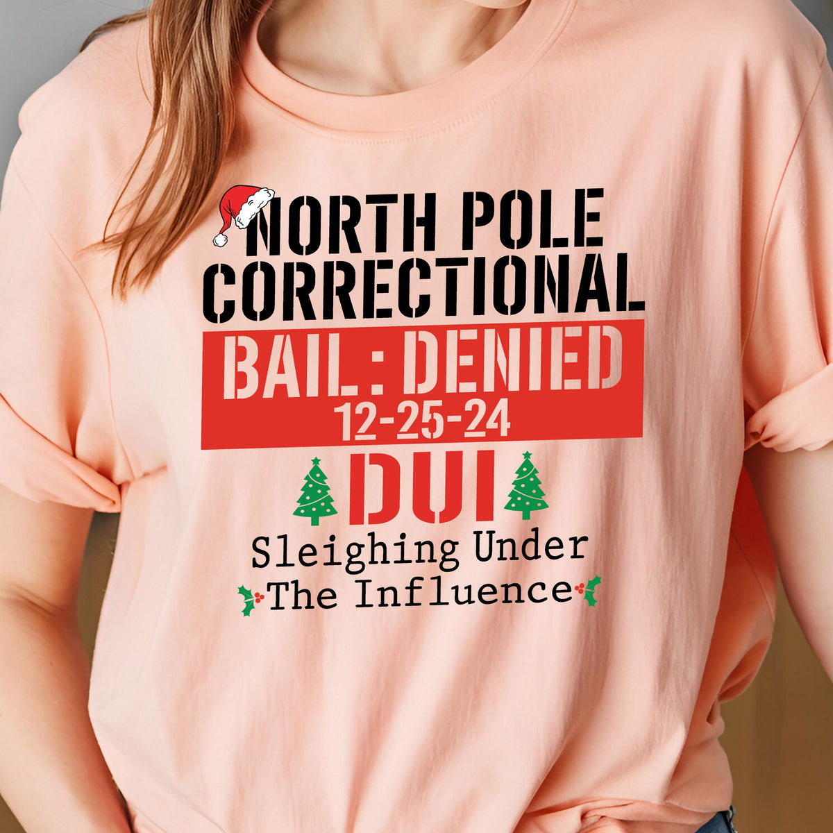 May include: A peach-colored t-shirt featuring black, red, and green text that reads NORTH POLE CORRECTIONAL BAIL: DENIED 12-25-24 DUI Sleighing Under The Influence, accented with a small red Santa hat and two green Christmas tree graphics.
