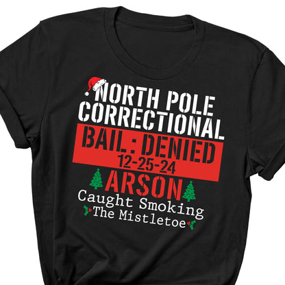 May include: Black t-shirt with white text that reads "NORTH POLE CORRECTIONAL BAIL: DENIED 12-25-24 ARSON Caught Smoking The Mistletoe" in a stencil font. A Santa hat graphic is above the text.