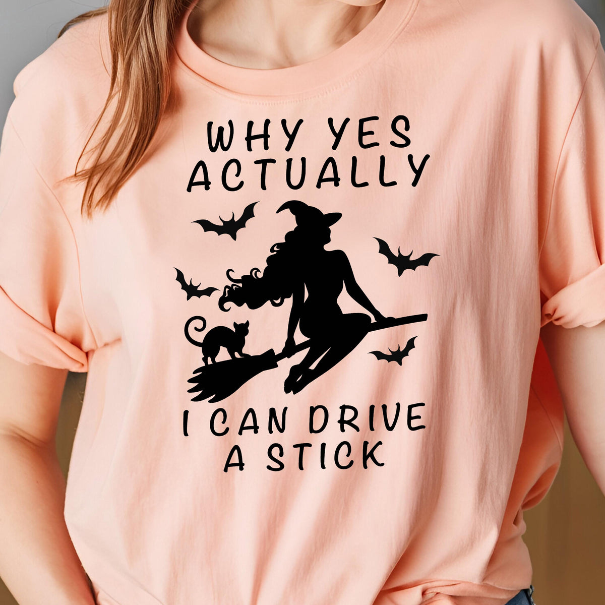 May include: A peach-colored t-shirt featuring a black silhouette of a witch riding a broom, with a black cat and bats. The text "WHY YES ACTUALLY" is above the image, and "I CAN DRIVE A STICK" is below.