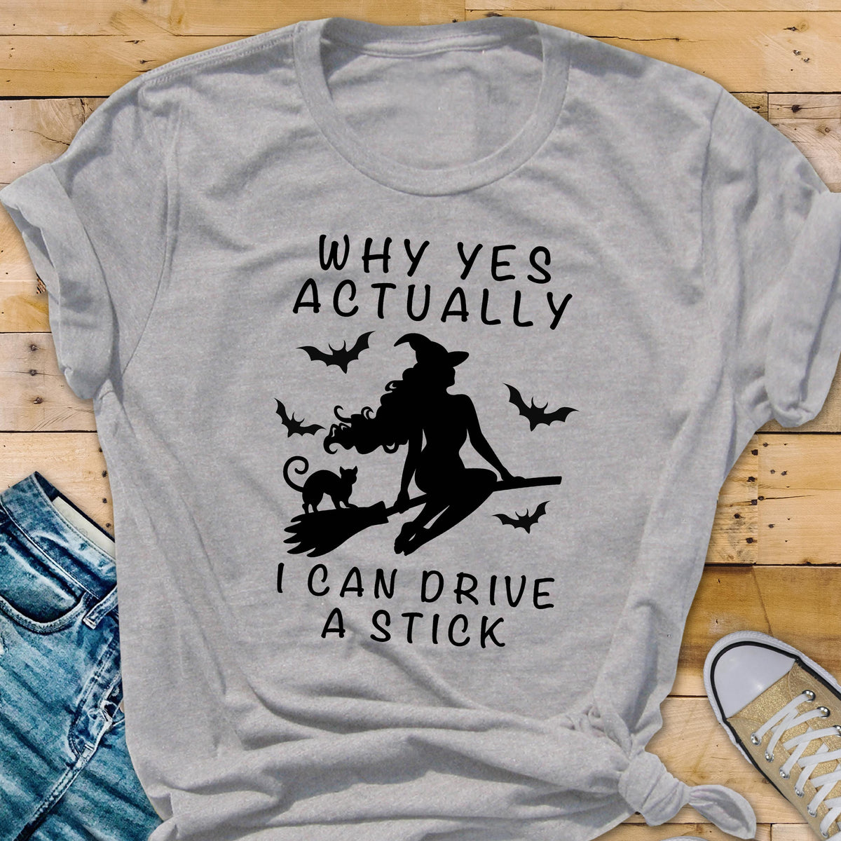 May include: A heather gray t-shirt featuring a black silhouette of a witch riding a broom, accompanied by a black cat and bats. The text on the shirt reads "WHY YES ACTUALLY I CAN DRIVE A STICK."