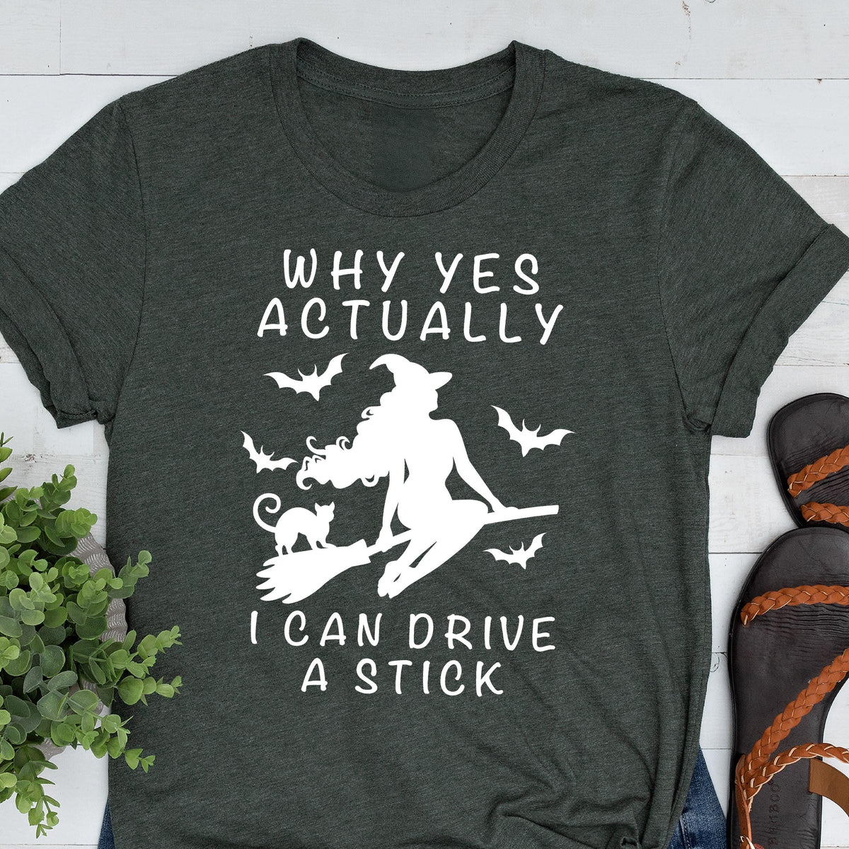 May include: Dark gray t-shirt with white text and graphic. The text reads "WHY YES ACTUALLY" above a silhouette of a witch on a broom with bats, and the text "I CAN DRIVE A STICK" below. The shirt has rolled-up sleeves.