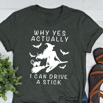 May include: Dark gray t-shirt with white text and graphic. The text reads "WHY YES ACTUALLY" above a silhouette of a witch on a broom with bats, and the text "I CAN DRIVE A STICK" below. The shirt has rolled-up sleeves.