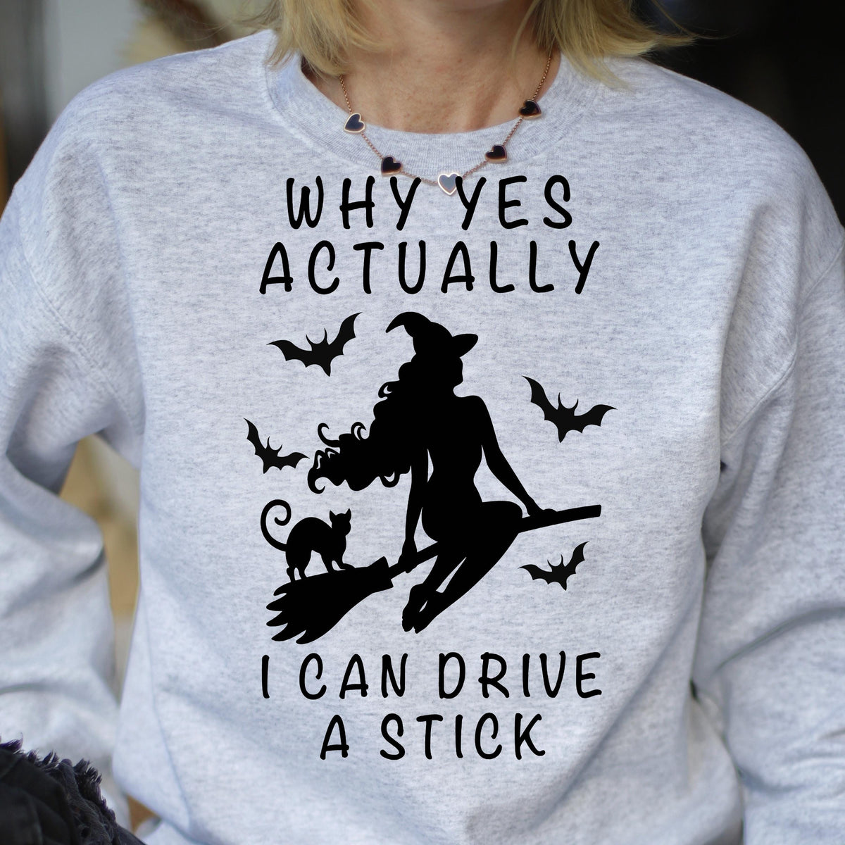 May include: A light gray crewneck sweatshirt featuring a black graphic of a witch riding a broomstick with a cat and bats. The text reads: WHY YES ACTUALLY I CAN DRIVE A STICK. The design is centered on the chest of the long-sleeved pullover garment.