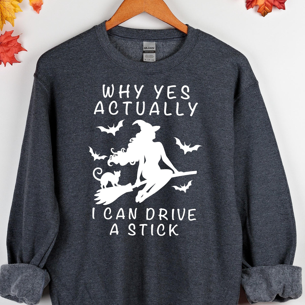 May include: A dark heather gray long-sleeve sweatshirt featuring a white graphic of a witch riding a broomstick with a cat and bats. The text reads: WHY YES ACTUALLY I CAN DRIVE A STICK. The garment has a crew neckline and ribbed cuffs.