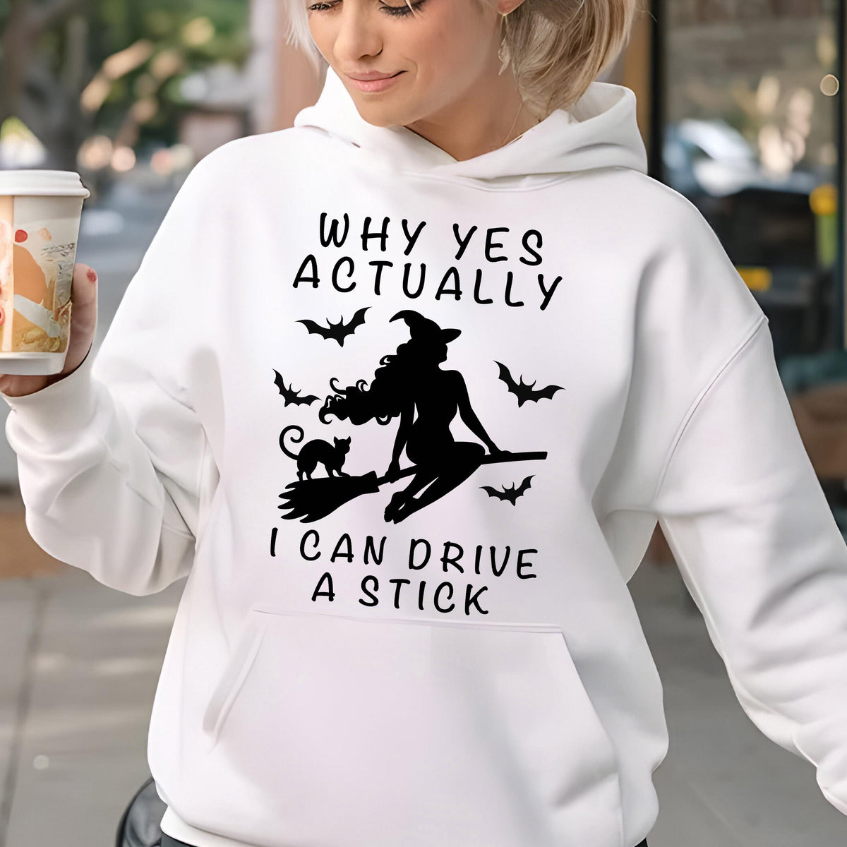 May include: A white hooded sweatshirt featuring a black graphic print of a witch riding a broomstick with a cat and bats. The text reads: WHY YES ACTUALLY I CAN DRIVE A STICK. The garment includes a front kangaroo pocket and long sleeves.