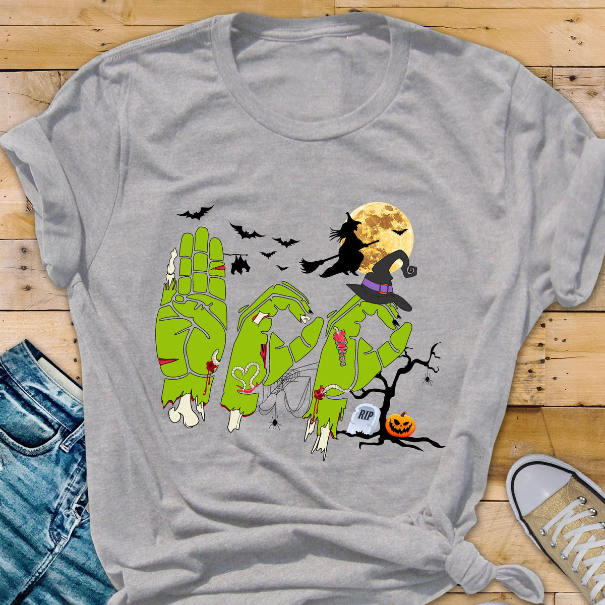 May include: A light gray t-shirt featuring a graphic of three green zombie hands forming the word BOO. The design includes a witch on a broomstick, a full moon, bats, a spider web, a bare tree, a tombstone with RIP text, and a jack-o'-lantern.