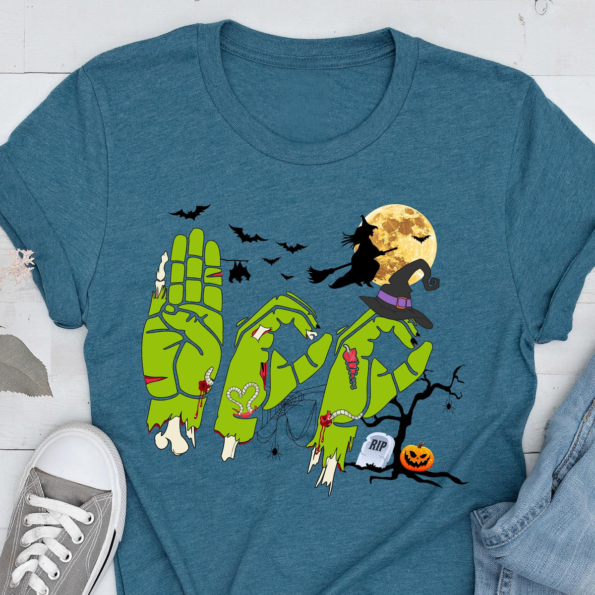 May include: A teal short-sleeve t-shirt featuring a graphic of three green zombie hands forming the letters B-O-O. The design includes a witch on a broomstick, a full moon, bats, a spider web, a tombstone with RIP text, and a jack-o'-lantern.