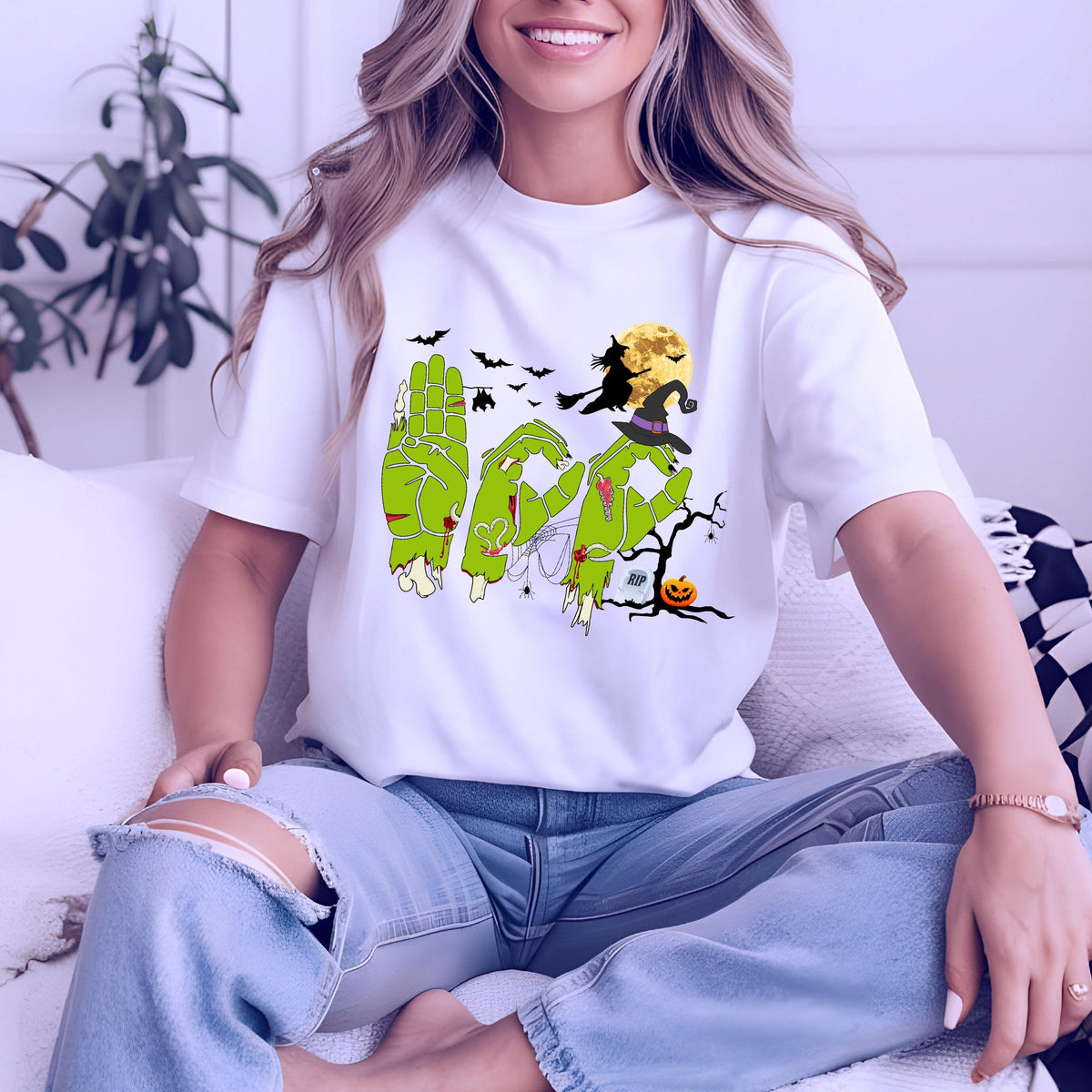 May include: A white short-sleeve t-shirt featuring a graphic of green zombie hands forming the word BOO in American Sign Language. The design includes a witch on a broomstick, a full moon, bats, a tombstone, a jack-o'-lantern, and a bare tree.