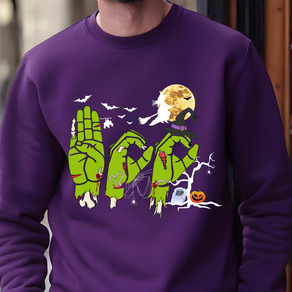 May include: A purple sweatshirt with a Halloween-themed design. The design features green zombie hands spelling out the word "BOO", bats, a full moon, a witch, a tombstone, and a jack-o'-lantern. The sweatshirt is made of a soft material.