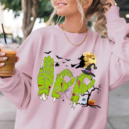 May include: A light pink sweatshirt with a Halloween-themed graphic. The graphic features green zombie hands spelling out the word "Boo", a witch flying on a broom, bats, a full moon, and a jack-o'-lantern. The sweatshirt has a crew neck and long sleeves.