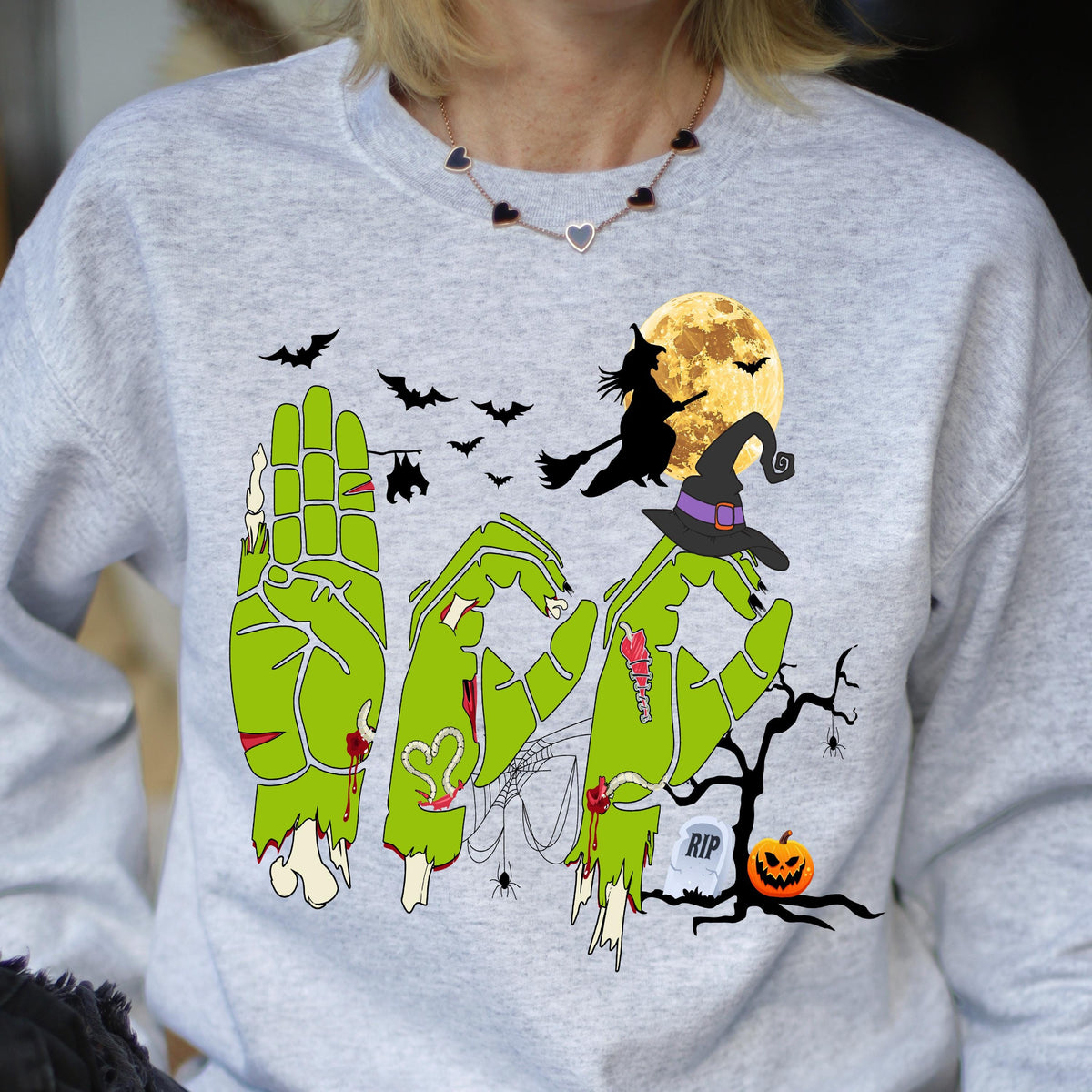 May include: A light gray sweatshirt with a Halloween-themed graphic. The design features green zombie hands spelling out the word "BOO", a witch on a broom, bats, a full moon, a witch's hat, a tombstone, and a jack-o'-lantern.