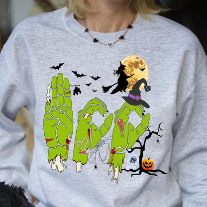 May include: A light gray sweatshirt with a Halloween-themed graphic. The design features green zombie hands spelling out the word "BOO", a witch on a broom, bats, a full moon, a witch's hat, a tombstone, and a jack-o'-lantern.