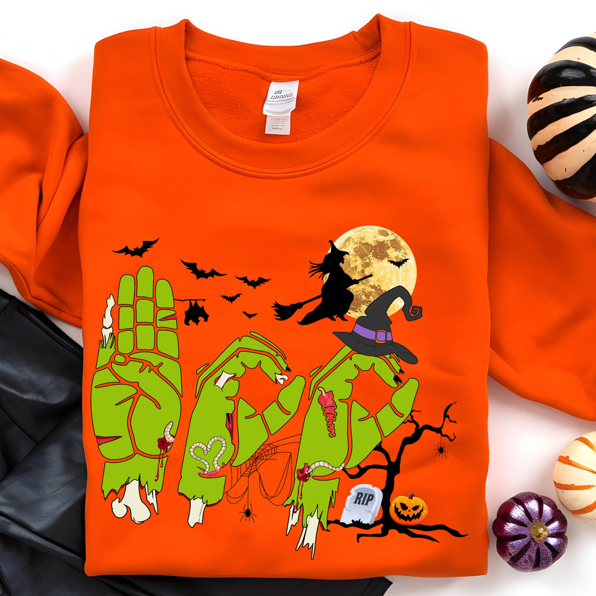 May include: Orange Halloween-themed sweatshirt with a green zombie hand design spelling out the word "BOO". The design includes a witch on a broom, bats, a full moon, and a tombstone. A spooky pumpkin and other Halloween decorations are also visible.